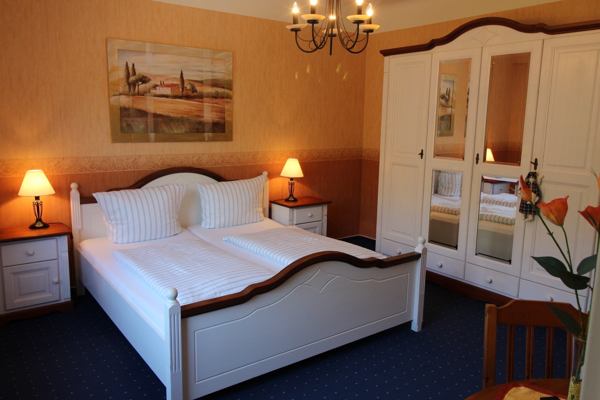 Zimmer ‚Milano‘ in der Pension Rehwinkel in Nalbach, mit elegantem weißen Bettgestell, passendem Kleiderschrank, warmen Wandfarben, kunstvollem Landschaftsgemälde und einem einladenden Kronleuchter.